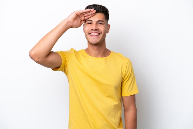 Junger kaukasischer Mann lokalisiert auf weißem Hintergrund, der mit der Hand mit glücklichem Ausdruck salutiert