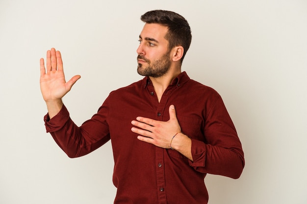 Junger kaukasischer Mann lokalisiert auf weißer Wand, die einen Eid leistet und Hand auf Brust legt.