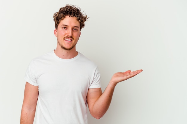 Junger kaukasischer Mann lokalisiert auf weißer Wand, die einen Kopienraum auf einer Handfläche zeigt und eine andere Hand auf Taille hält.