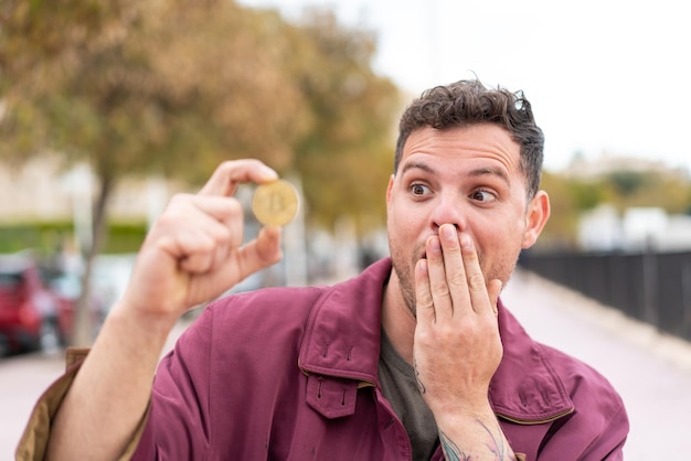 Junger kaukasischer Mann mit einem Bitcoin im Freien mit Überraschung und schockiertem Gesichtsausdruck