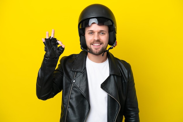 Junger kaukasischer Mann mit einem Motorradhelm auf gelbem Hintergrund isoliert, der ein OK-Zeichen mit den Fingern zeigt