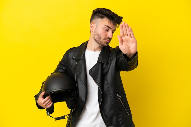 Junger kaukasischer Mann mit einem Motorradhelm auf gelbem Hintergrund isoliert, der eine Stopp-Geste macht und enttäuscht