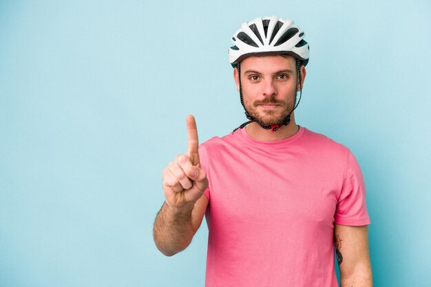 Junger kaukasischer mann mit fahrradhelm lokalisiert auf blauem hintergrund, der nummer eins mit dem finger zeigt