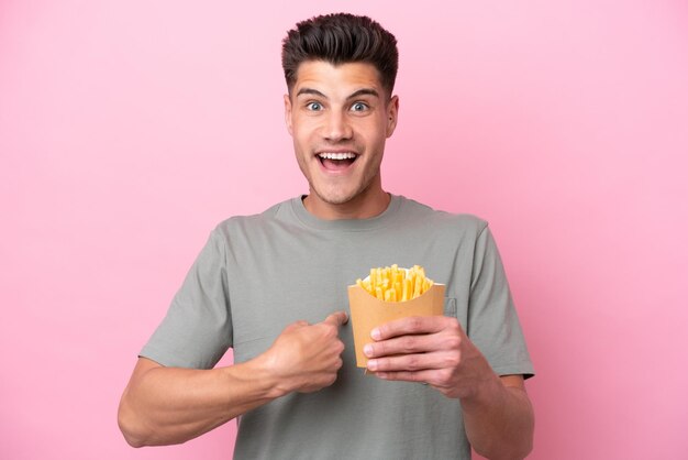 Junger kaukasischer Mann mit gebratenen Chips isoliert auf rosa Hintergrund mit überraschendem Gesichtsausdruck