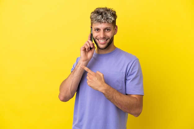 Junger kaukasischer Mann mit Handy isoliert auf gelbem Hintergrund, der auf die Seite zeigt, um ein Produkt zu präsentieren