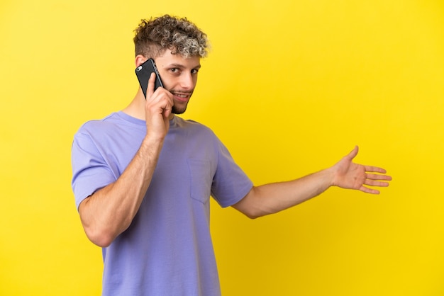 Junger kaukasischer Mann mit Handy isoliert auf gelbem Hintergrund, der die Hände zur Seite ausstreckt, um einzuladen, zu kommen