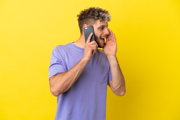 Junger kaukasischer Mann mit Handy isoliert auf gelbem Hintergrund und schreit mit weit geöffnetem Mund zur Seite