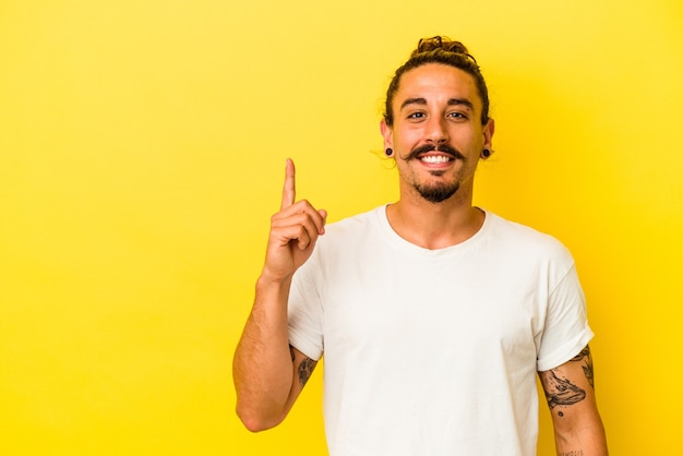 Junger kaukasischer Mann mit langen Haaren isoliert auf gelbem Hintergrund, der Nummer eins mit dem Finger zeigt.