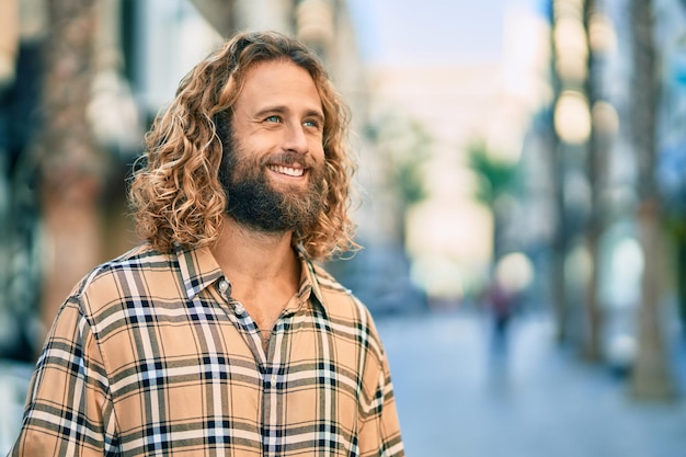 Junger kaukasischer Mann mit langen Haaren lächelt glücklich in der Stadt.