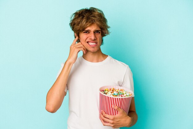 Junger kaukasischer Mann mit Make-up, das Popcorn isoliert auf blauem Hintergrund hält und die Ohren mit den Händen bedeckt.