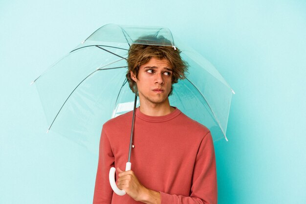 Junger kaukasischer Mann mit Make-up, der Regenschirm einzeln auf blauem Hintergrund hält, verwirrt, fühlt sich zweifelhaft und unsicher.