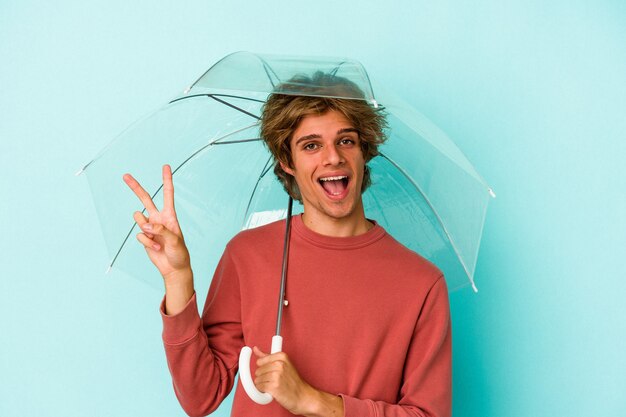 Junger kaukasischer Mann mit Make-up, der Regenschirm isoliert auf blauem Hintergrund hält, freudig und sorglos, der ein Friedenssymbol mit den Fingern zeigt.