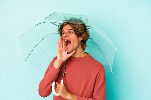 Junger kaukasischer Mann mit Make-up, der Regenschirm isoliert auf blauem Hintergrund hält und die Handfläche in der Nähe des geöffneten Mundes schreit und hält.