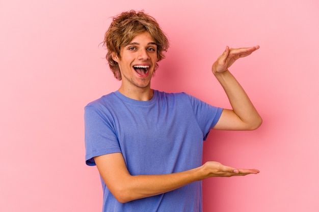 Junger kaukasischer Mann mit Make-up einzeln auf rosafarbenem Hintergrund, der etwas Kleines mit den Zeigefingern hält, lächelt und selbstbewusst.