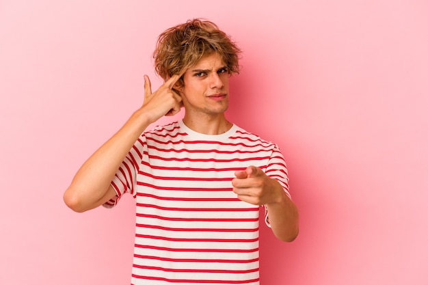 Junger kaukasischer Mann mit Make-up einzeln auf rosafarbenem Hintergrund, der mit dem Finger auf den Tempel zeigt, denkt, konzentriert sich auf eine Aufgabe.