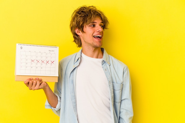 Junger kaukasischer Mann mit Make-up-Holding-Kalender einzeln auf gelbem Hintergrund sieht beiseite lächelnd, fröhlich und angenehm aus.