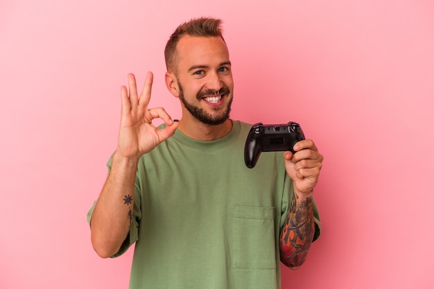 Junger kaukasischer Mann mit Tätowierungen, der einen Gamecontroller einzeln auf rosafarbenem Hintergrund hält, fröhlich und selbstbewusst, der eine ok Geste zeigt.