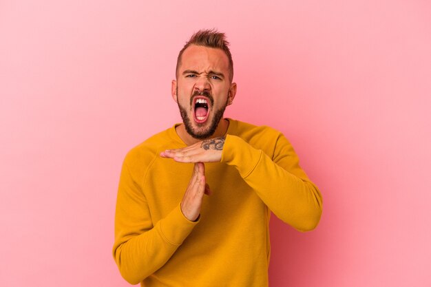 Junger kaukasischer Mann mit Tätowierungen, die auf rosafarbenem Hintergrund isoliert sind und eine Timeout-Geste zeigen.