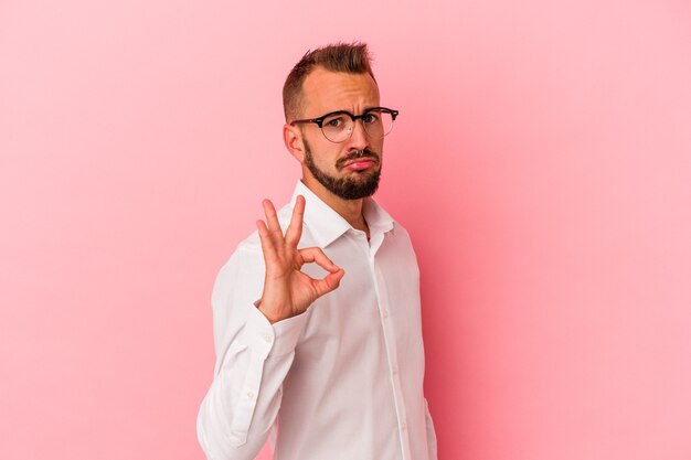 Foto junger kaukasischer mann mit tätowierungen, die auf rosafarbenem hintergrund isoliert sind, zwinkert ein auge und hält eine okaygeste mit der hand.