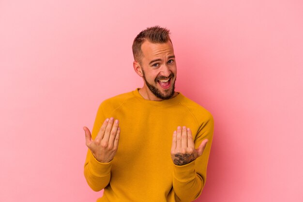 Junger kaukasischer Mann mit Tätowierungen einzeln auf rosafarbenem Hintergrund, der mit dem Finger auf Sie zeigt, als ob er einladen würde, näher zu kommen.