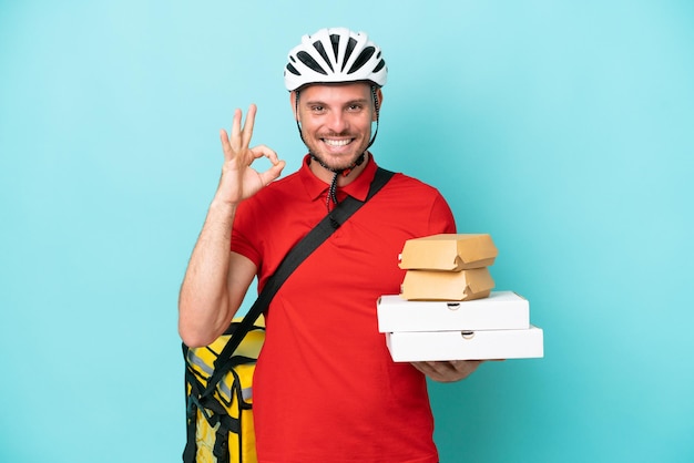 Junger kaukasischer Mann mit Thermorucksack und Fast Food isoliert auf blauem Hintergrund, der mit den Fingern ein Ok-Zeichen zeigt