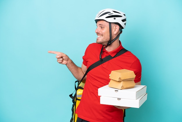 Junger kaukasischer Mann mit Thermorucksack und hält Fast Food isoliert auf blauem Hintergrund, zeigt mit dem Finger zur Seite und präsentiert ein Produkt