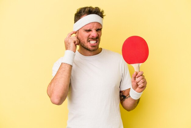 Junger kaukasischer Mann mit Tischtennisschläger isoliert auf gelbem Hintergrund, der die Ohren mit den Händen bedeckt.