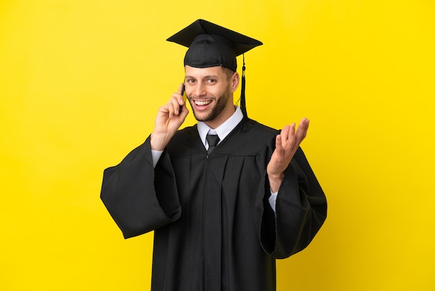 Junger kaukasischer Mann mit Universitätsabschluss isoliert auf gelbem Hintergrund, der ein Gespräch mit dem Handy mit jemandem führt