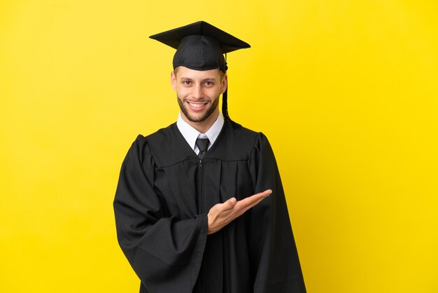 Junger kaukasischer Mann mit Universitätsabschluss isoliert auf gelbem Hintergrund, der eine Idee präsentiert, während er lächelnd in Richtung blickt