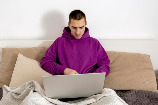 Junger kaukasischer mann mit violettem kapuzenpulli, der auf bett sitzt und laptop-computer hält. Mann, der ein Notebook verwendet, um im Internet zu surfen, Nachrichten zu lesen, Filme anzusehen, zu lernen oder online zu arbeiten. Zu Hause entspannen.