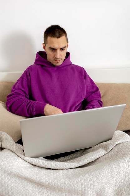 Junger kaukasischer mann mit violettem kapuzenpulli, der auf bett sitzt und laptop-computer hält. Mann, der ein Notebook verwendet, um im Internet zu surfen, Nachrichten zu lesen, Filme anzusehen, zu lernen oder online zu arbeiten. Zu Hause entspannen.