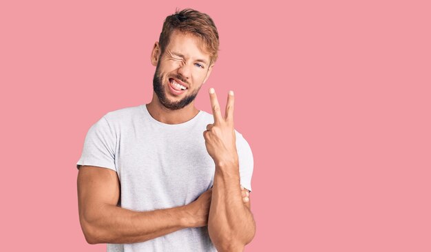 Junger kaukasischer Mann trägt lässiges weißes T-Shirt, lächelt mit glücklichem Gesicht und zwinkert in die Kamera und macht ein Siegeszeichen. Nummer zwei.