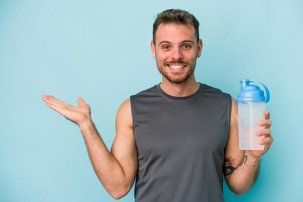 Junger kaukasischer Mann trinkt Milchshake isoliert auf blauem Hintergrund, zeigt einen Kopierraum auf einer Handfläche und hält eine andere Hand an der Taille