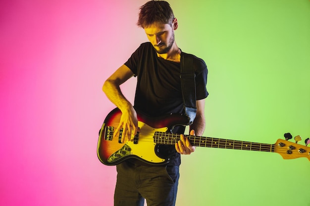 Junger kaukasischer Musiker, der Bassgitarre im Neonlicht auf rosa-grünem Hintergrund spielt