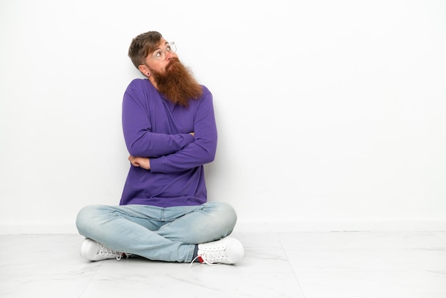 Junger kaukasischer rötlicher Mann, der auf dem Boden sitzt, isoliert auf weißem Hintergrund, der beim Anheben der Schultern Zweifel macht