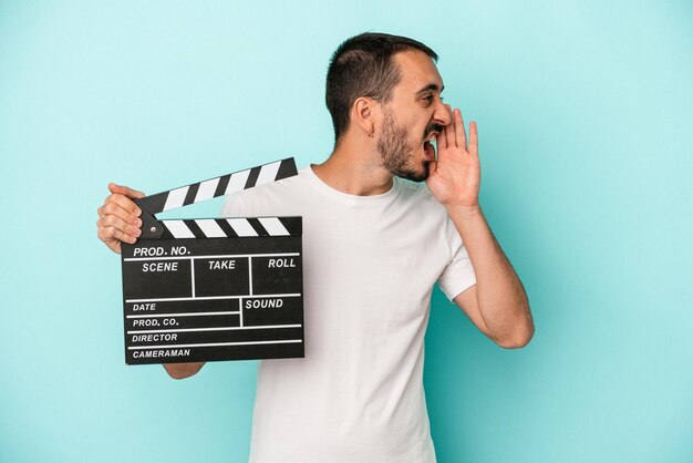 Junger kaukasischer Schauspieler, der die Klappe isoliert auf blauem Hintergrund hält, schreit und die Handfläche in der Nähe des geöffneten Mundes hält