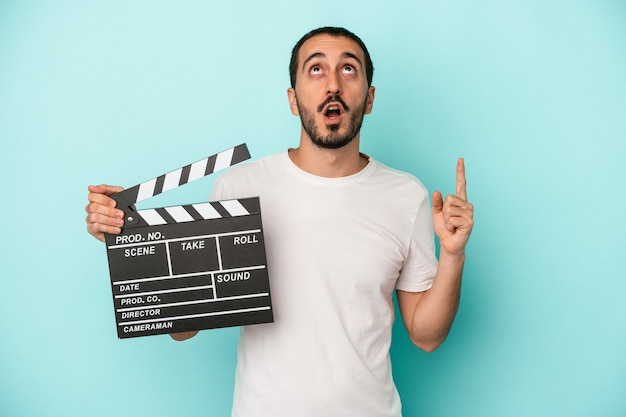 Junger kaukasischer Schauspielermann, der clapperboard lokalisiert auf blauem Hintergrund hält, der mit geöffnetem Mund nach oben zeigt.