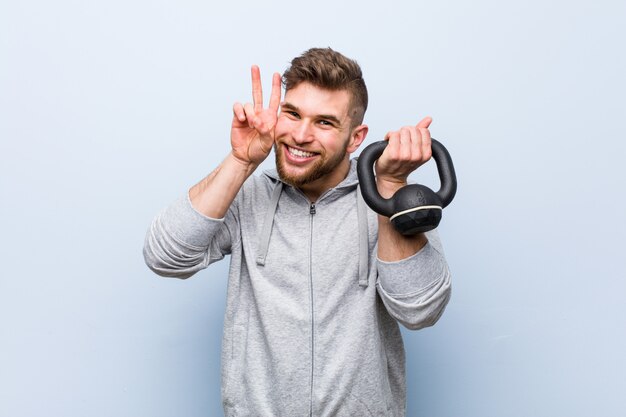 Junger kaukasischer Sportler, der eine Hantel hält, die Siegeszeichen zeigt und breit lächelt.