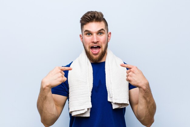 Junger kaukasischer Sportler mit einem Handtuch überrascht auf sich selbst zeigend, breit lächelnd.