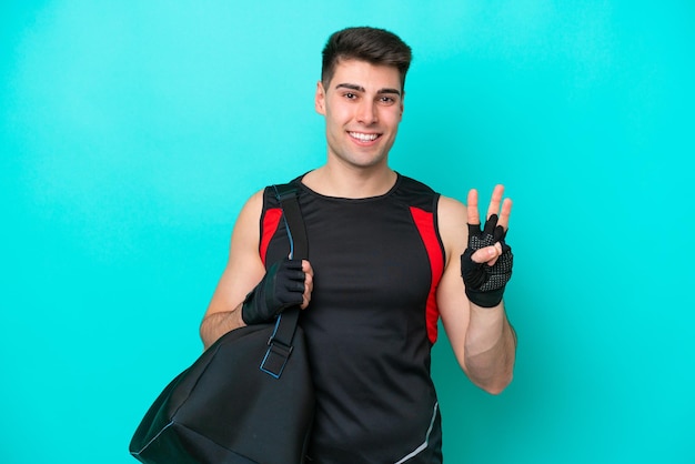 Junger kaukasischer Sportler mit Sporttasche isoliert auf blauem Hintergrund glücklich und zählt drei mit den Fingern