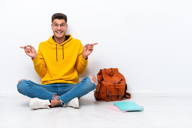 Junger kaukasischer Student, der auf dem Boden sitzt, isoliert auf weißem Hintergrund, zeigt mit dem Finger auf die Seitenteile und ist glücklich