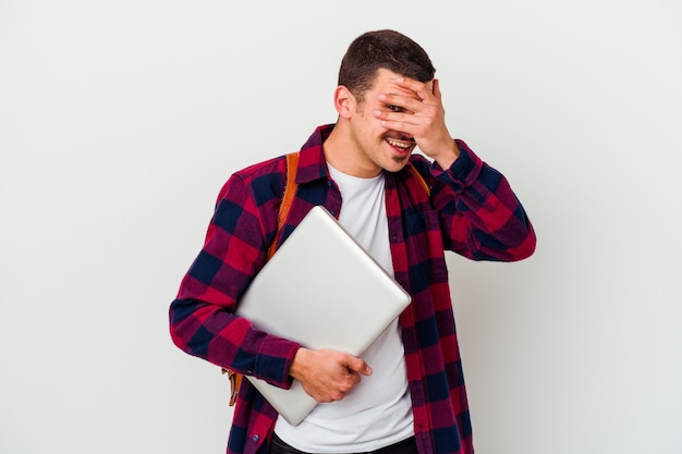 Junger kaukasischer Studentenmann, der einen Laptop lokalisiert auf weißem Blinzeln an der Kamera durch Finger hält, verlegenes bedeckendes Gesicht.