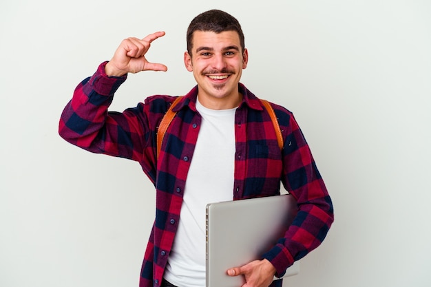 Junger kaukasischer Studentenmann, der einen Laptop lokalisiert auf weißer Wand hält, die etwas wenig mit Zeigefingern hält, lächelnd und zuversichtlich.