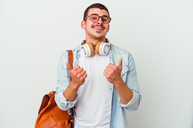 Junger kaukasischer Studentenmann, der Musik auf Weiß hört, der beide Daumen anhebt, lächelnd und zuversichtlich.