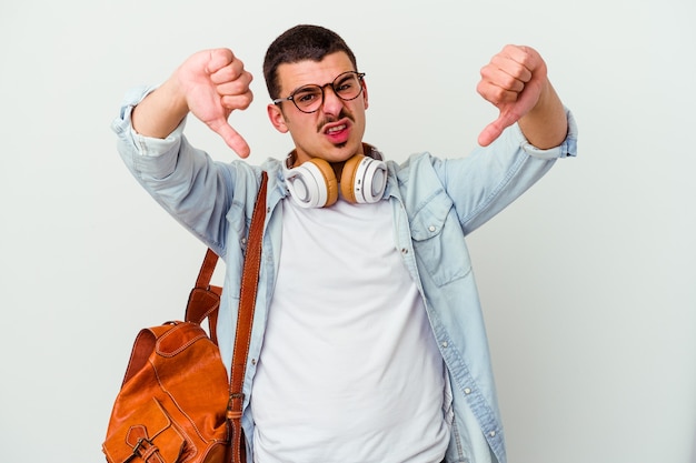 Junger kaukasischer studentenmann, der musik hört, die auf weiß lokalisiert wird, das daumen nach unten zeigt und abneigung ausdrückt.