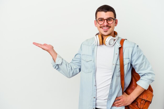 Junger kaukasischer Studentenmann, der Musik lokalisiert auf weißer Wand hört, die einen Kopienraum auf einer Handfläche zeigt und eine andere Hand auf Taille hält.