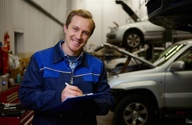 Junger kaukasischer techniker lächelt in die kamera und macht checkliste für die autoreparatur in der garage. Autoservice, Reparatur, Autogarantie-Servicekonzept, Automechaniker schreibt auf einem Klemmbrett in einer Werkstatt.