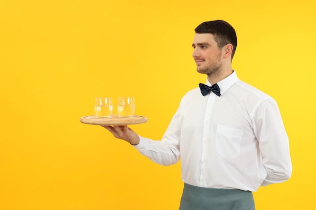 Junger Kellner hält Tablett mit Gläsern Wasser auf gelbem Hintergrund.