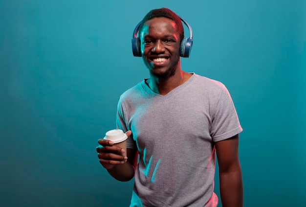 Junger Kerl, der eine Tasse Kaffee hält und Musik auf dem Headset genießt, zeigt ein offenes Lächeln vor der Kamera. Zuversichtliches Modell, das drahtlose Kopfhörer verwendet, um Aufnahmelieder zu hören, während er ein Getränk in der Hand hat.