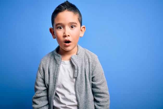 Junger kleiner Junge trägt Sport-Sweatshirt vor blauem, isoliertem Hintergrund, ängstlich und schockiert mit überraschendem Gesichtsausdruck, Angst und aufgeregtem Gesicht